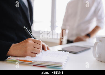 Close up view of woman taking notes with male colleague in background. Focus on hands of businesswoman. Stock Photo