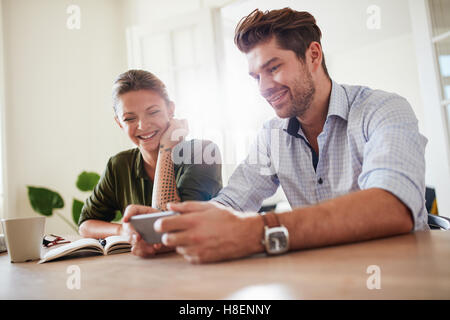 Shot of happy young couple looking at smart phone and smiling. Handsome young man with beautiful girlfriend sitting at table usi Stock Photo
