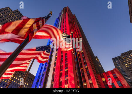 Rockefeller Center at twilight illuminated in white, red and blue. American flags flap in the wind. Midtown Manhattan, New York