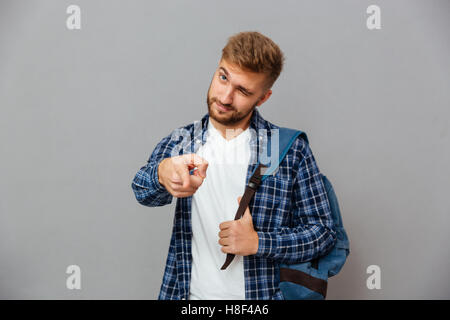 Portrait of a handsome bearded man with backpack pointing finger at camera and winking isolated on a gray background Stock Photo