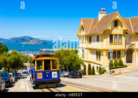 Blue cable car with hanging tourists going uphill on steep Hyde St with sweeping view of Alcatraz Prison, bay water and yellow V