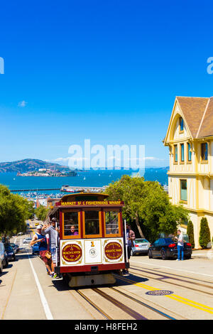 Tourists riding outside cable car climbing steep uphill street on Hyde St with view of Alcatraz Prison and Victorian house on bl