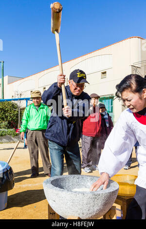 Japan. Omochi, winter rice bashing festival. Senior man holding with both hands a wooden mallet, a kine, beating rice in usu, bowl. Outdoors. Stock Photo