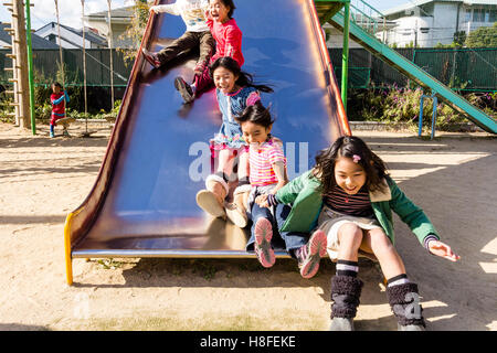 Japan. School playground. Children, three girls, 6-8 years old, jumping off slide in row, excited, shouting happily. Two boys sliding down behind them Stock Photo