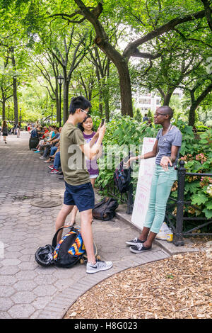 young Asian American couple in park aim smart phone as cool bald black woman barker pitchs website for crowdfound friends Stock Photo