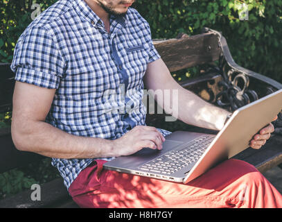 Young Man wearing in shirt sitting in city park and writing on his laptop, modern design notebook Stock Photo