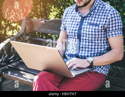 Young Man wearing in shirt sitting in city park and writing on his laptop, watch on wrist Stock Photo