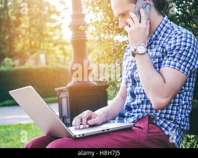 Young man talking by smartphone with laptop on his knees Stock Photo