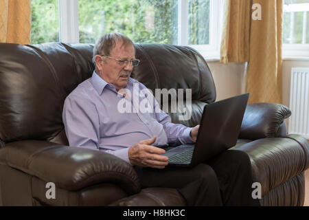 Elderly man getting frustrated by a laptop, he is looking confused whilst sat on the sofa Stock Photo