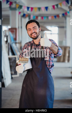 Smiling house painting worker equipped with protective suit and showing thumbs up. Workshop on a background Stock Photo