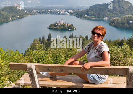 White middle aged tanned woman sit on wooden bench with view from above of Lake Bled. From mountain Osojnica in Slovenia. Stock Photo