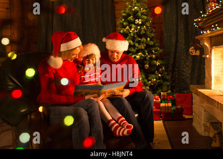 Kid with parents read stories sitting on coach in front of fireplace in Christmas decorated house interior