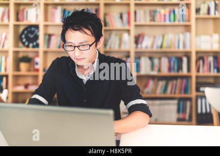 Young pensive Asian man working on laptop at home office or library with serious face, bookshelf blur background with copy space Stock Photo