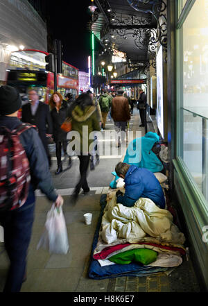 Homeless man and begging cup with his entire belongings living and sleeping on busy Shaftesbury Avenue, with bus, people, shoppers, blurred passing by at night London UK. Stock Photo