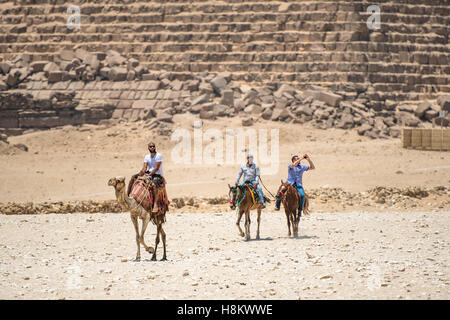 Cairo, Egypt Camel drivers on horseback and a tourist riding a camel walking through the desert with the Great Pyramids of Giza  Stock Photo