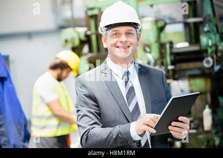 Portrait of confident mature businessman using digital tablet with worker in background at factory Stock Photo