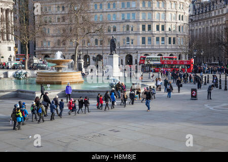A 'crocodile' of school children follow their teacher across Trafalgar Square with the fountains and red double decker buses Stock Photo
