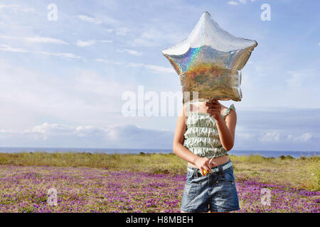 Front view of preadolescent girl holding star shaped balloon in front of her face while standing at flower field Stock Photo