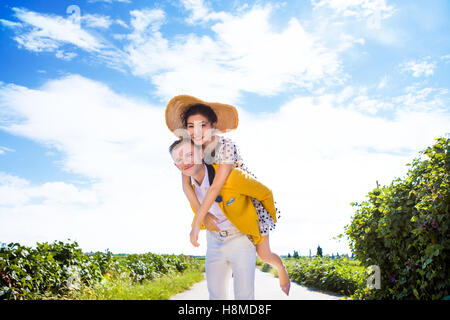 Portrait of happy man piggybacking woman on footpath amidst field against cloudy sky Stock Photo