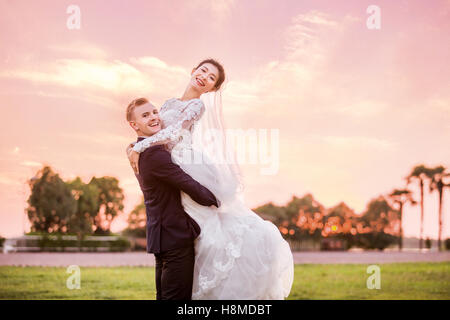 Side view portrait of happy bridegroom carrying bride on field during sunset Stock Photo