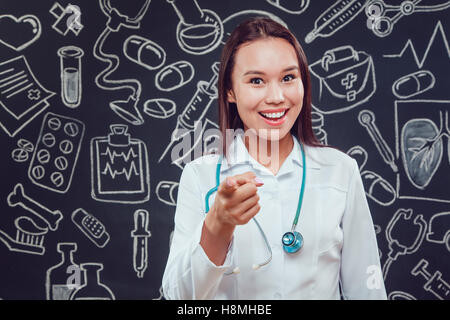 Smiling young woman doctor in white coat standing on dark background with pattern. He points the finger forward Stock Photo