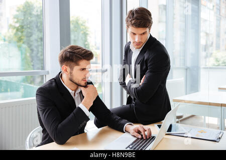 Two serious young businessmen working and using laptop in office together Stock Photo