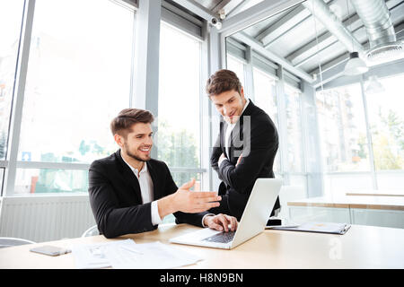 Two happy young businessmen talking and using laptop in office together Stock Photo