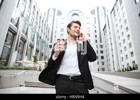 Happy young businessman talking on mobile phone and drinking coffee near business center Stock Photo