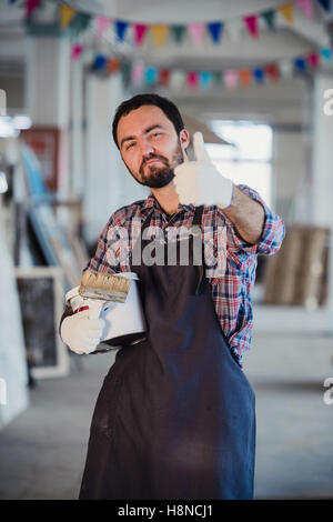 Smiling house painting worker equipped with protective suit and showing thumbs up. Workshop on a background Stock Photo