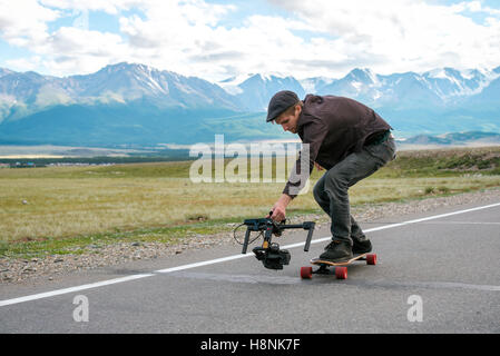 Man rides a longboard and shoot camera with electronic steadycam. Long road in the field on  background of mountains  glaciers. Stock Photo