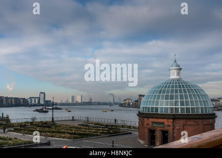 The domed entrance to Greenwich foot tunnel in the foreground with a view along the River Thames west towards the skyline of the Stock Photo