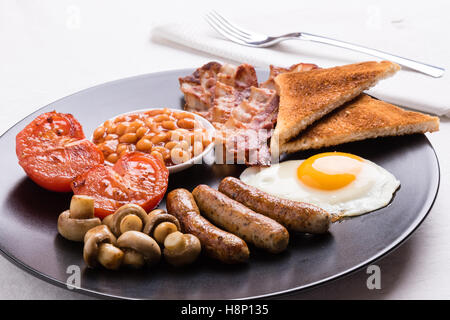 full english breakfast with bacon, sausage, eggs, beans, mushrooms on black plate Stock Photo