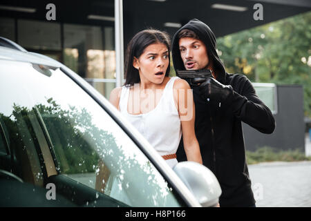 Aggressive man thief in gloves threatening with gun to frightened young woman on car parking Stock Photo