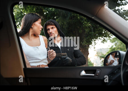 Dangerous criminal man with gun stealing car of scared young woman on outdoor parking Stock Photo