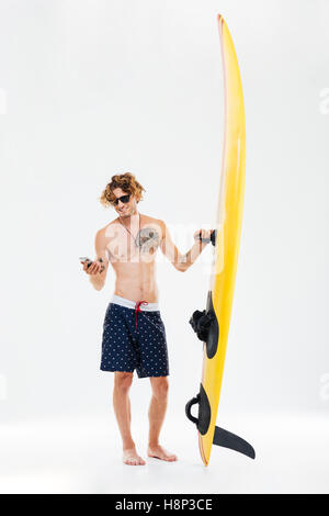 Happy young sportsman holding surfboard and using mobile phone isolated on the white background Stock Photo