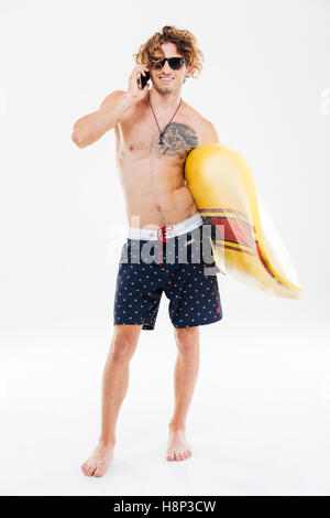 Full length portrait of a handsome young sportsman holding surfboard and talking on mobile phone isolated on the white backgroun Stock Photo