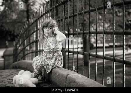 Cute little girl sat on a bridge pretending to talk on a mobile phone Stock Photo