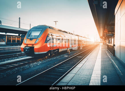 Modern high speed red commuter train at the railway station at colorful sunset. Railroad with vintage toning. Train at railway p Stock Photo