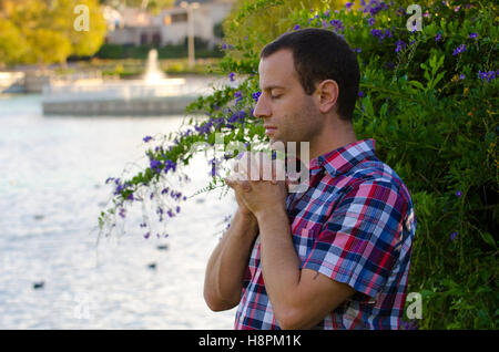 Man praying by a lake alone in front of a bush with flowers and a fountain in the background. Stock Photo