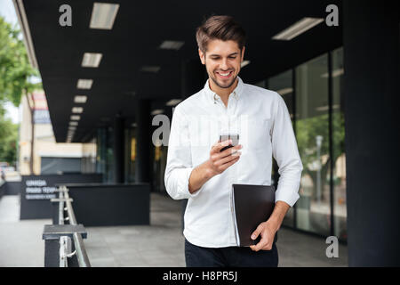 Happy attractive young businessman walking and using mobile phone outdoors Stock Photo