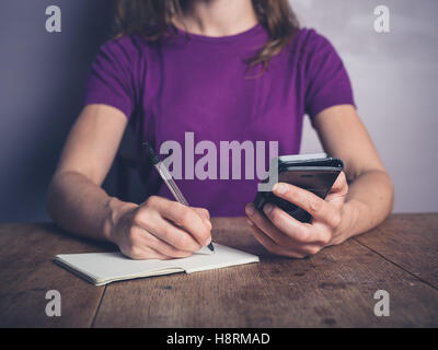 A young woman is sitting at a table using a smartphone and writing in a notepad Stock Photo