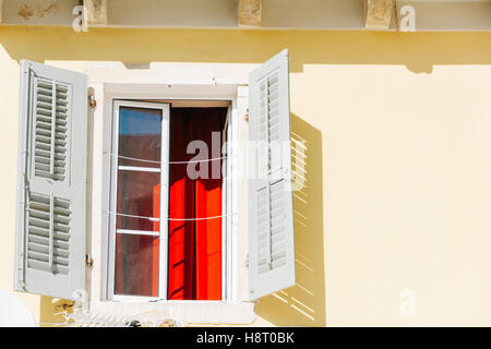 An open window with wooden shutters and red curtains on a sunny day Stock Photo