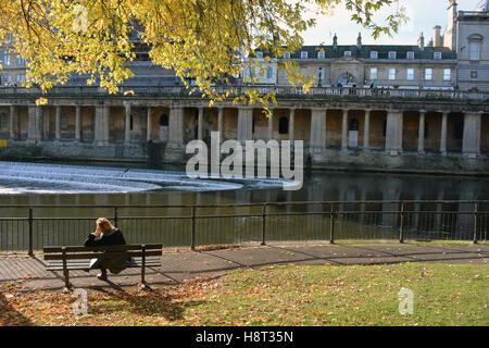 Woman on park bench on Riverside Walk looking across River Avon to Grand Parade, Bath, Somerset, England Stock Photo