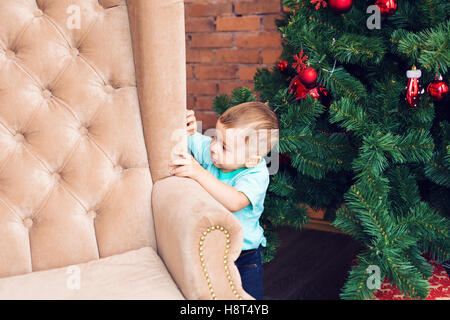 Funny little boy standing near sofa and Christmas tree with colorful xmas ball Stock Photo