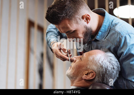 Handsome young barber cutting mustache of senior client with scissors Stock Photo