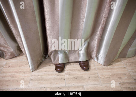 Shoes of a man hidden behind striped curtains in a room. Stock Photo