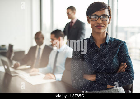 Confident young African businesswoman standing with folded arms smiling at the camera in a boardroom with male colleagues in the Stock Photo