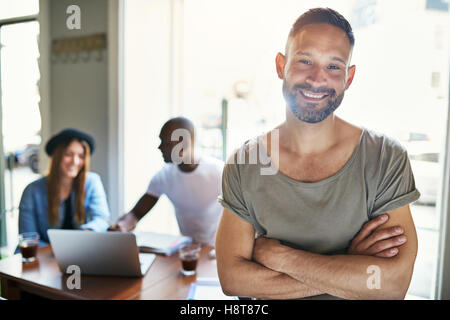 Handsome smiling bearded man in short sleeve shirt and folded arms standing in cafe with customers in background Stock Photo