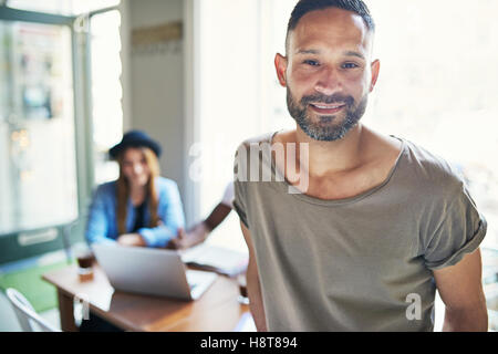 Handsome grinning bearded man in short sleeve shirt leaning on table in coffee house with woman in background Stock Photo