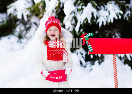 Happy child in knitted reindeer hat and scarf holding letter to Santa with Christmas presents wish list at red mail box in snow Stock Photo